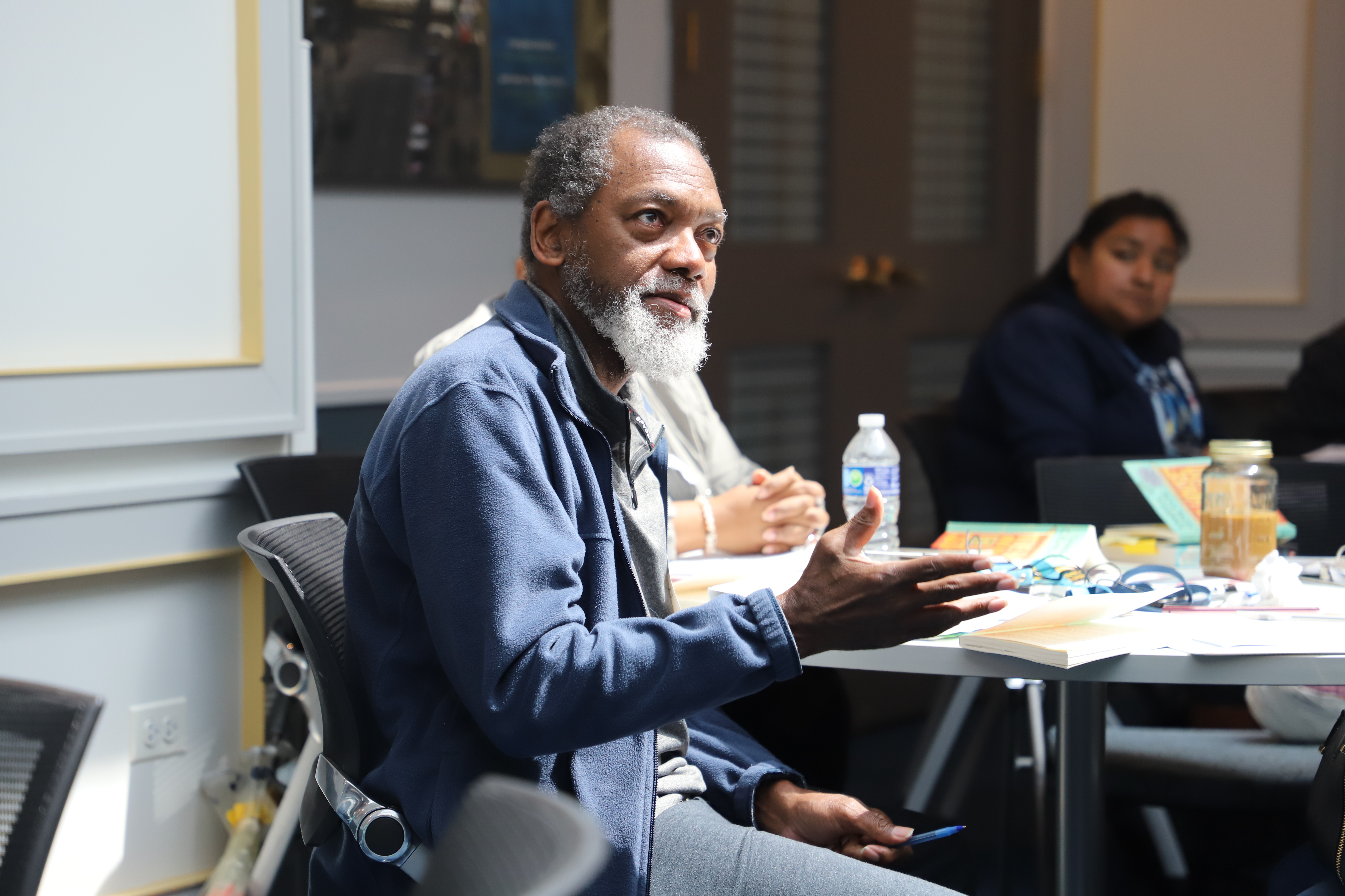 Participant speaking at a writing workshop, sitting down, surrounded by others.